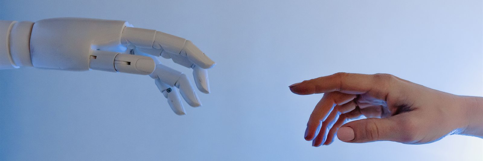 Fehér robotkéz és emberi kéz nyúl egymás felé világoskék háttér előtt