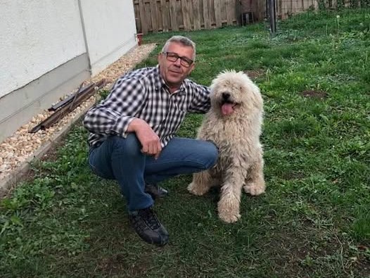 Egy férfi guggol a kertben egy kisebb termetű, fehér komondor mellett